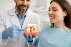 A dentist is pointing to a dental implant placed in a patient's mouth, using a model to show how dental implants for missing teeth work. No text on the image.